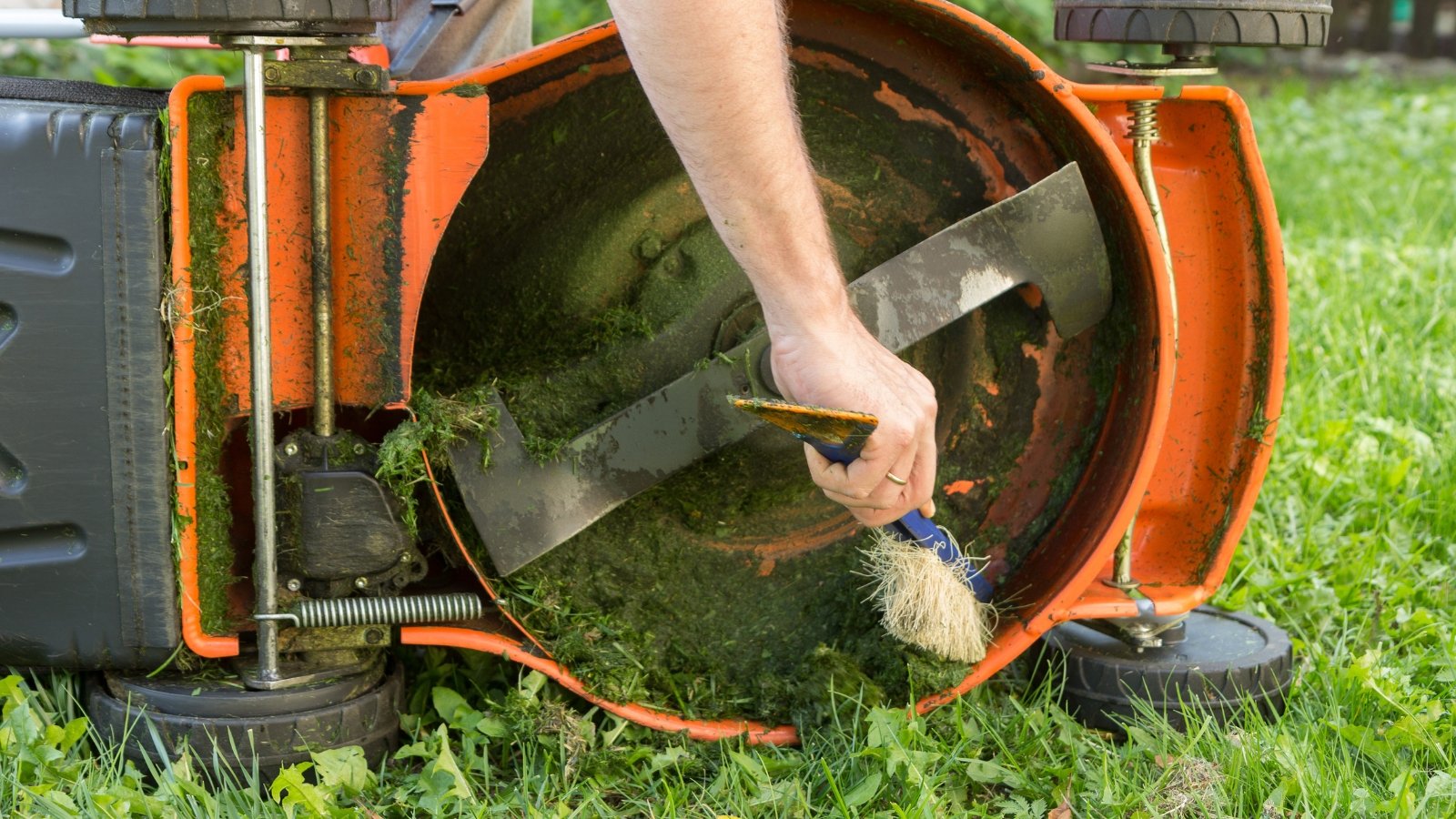 Hands reaching under a lawn mower to clear accumulated grass clippings and debris, focusing on the underside and blades to ensure clean operation.