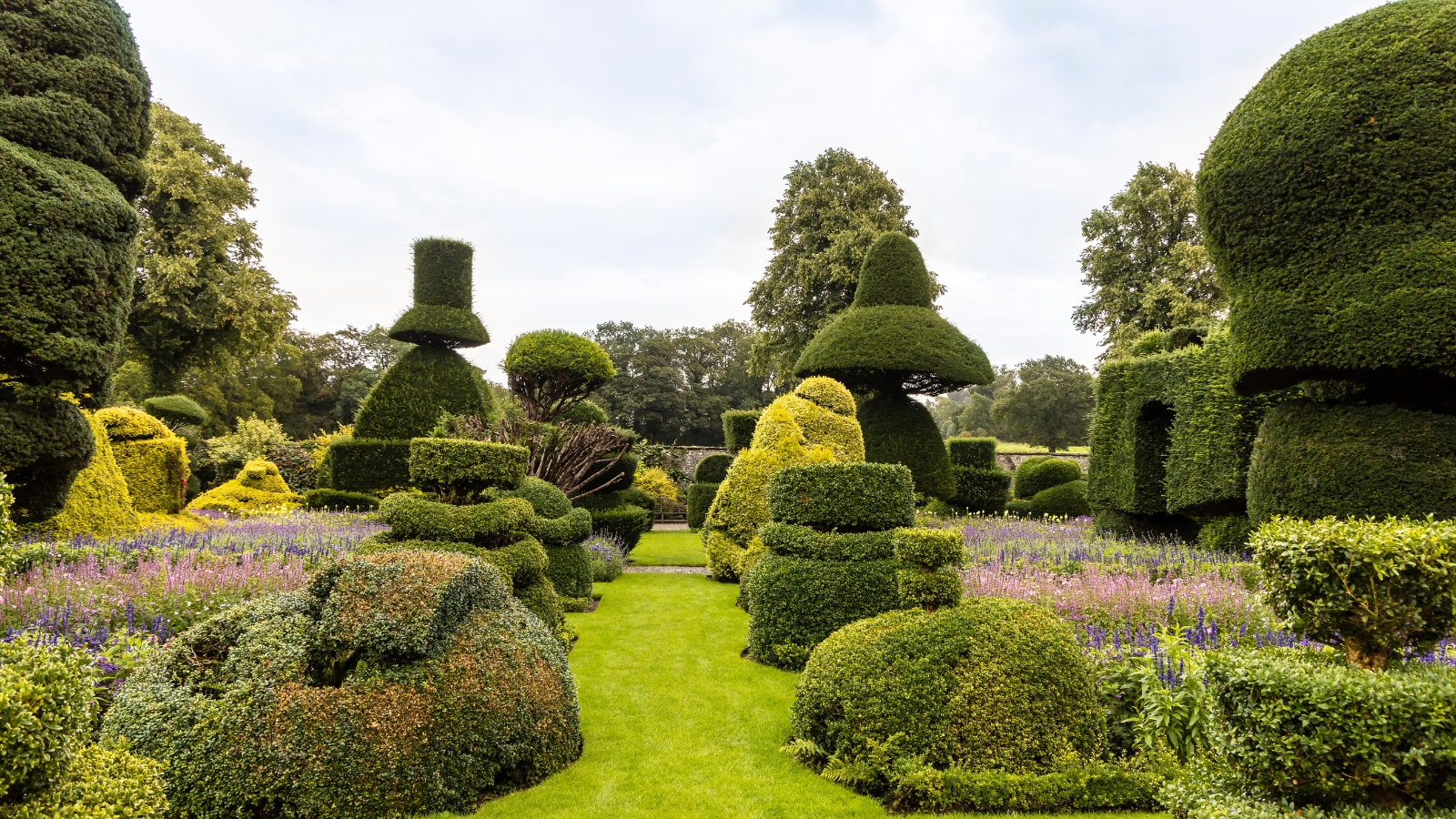 A topiary garden with intricate green sculptures shaped into cones, spheres, and other designs, surrounded by a manicured grass path and colorful flower beds.