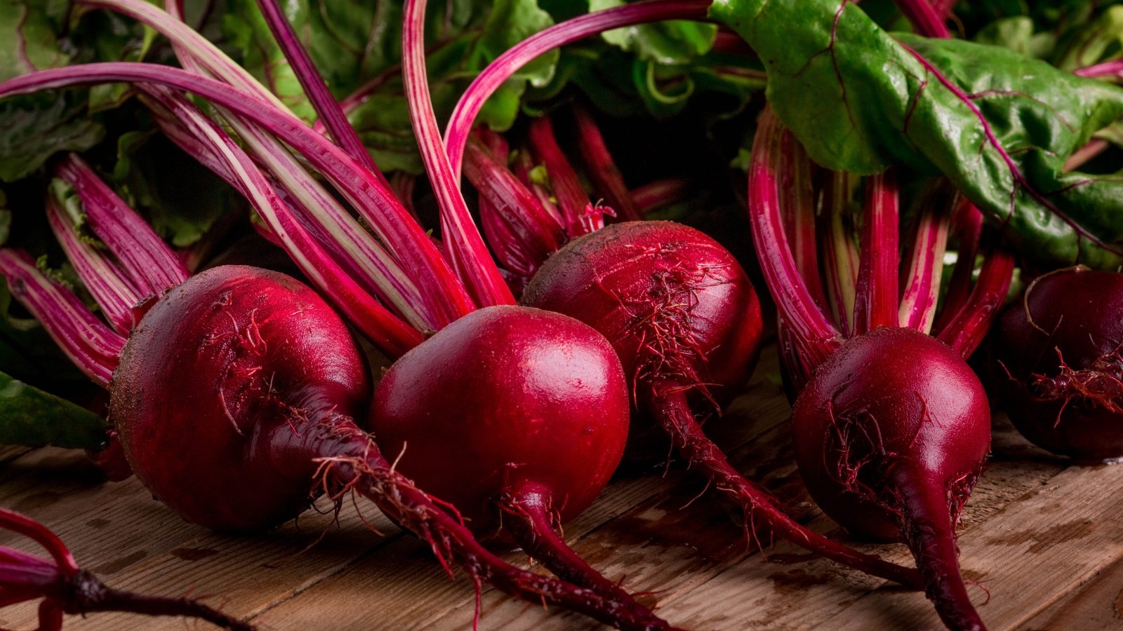 A collection of freshly pulled root vegetables with deep red skins and vibrant magenta stems, lying on a rustic wooden surface, with some soil remnants clinging to the roots.