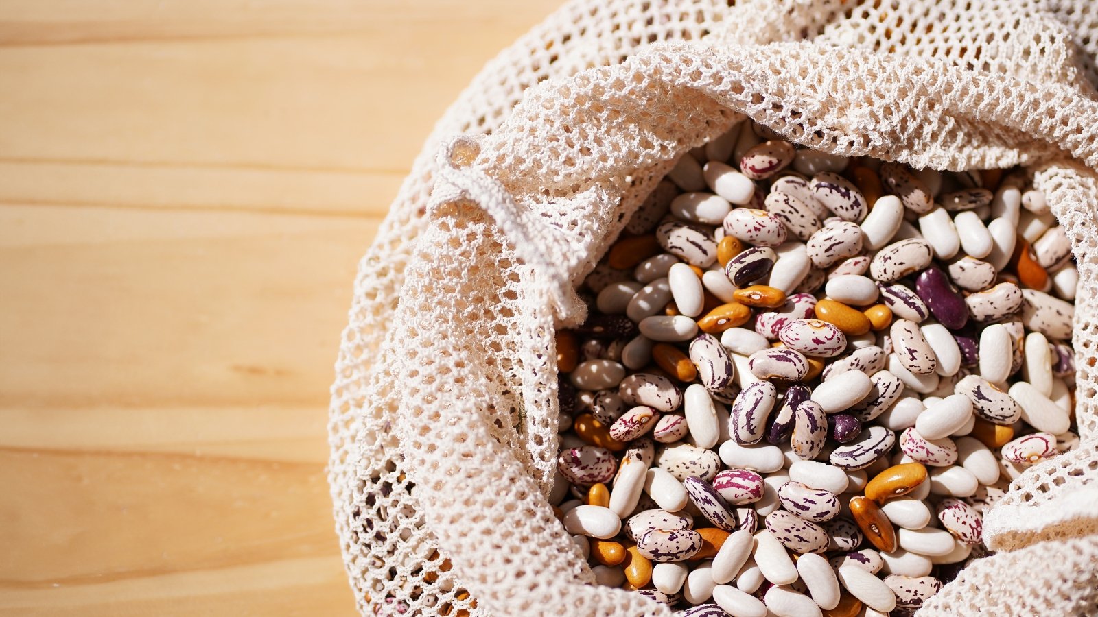 A crocheted reusable bag holds a colorful mix of speckled beans in shades of white, brown, and red.
