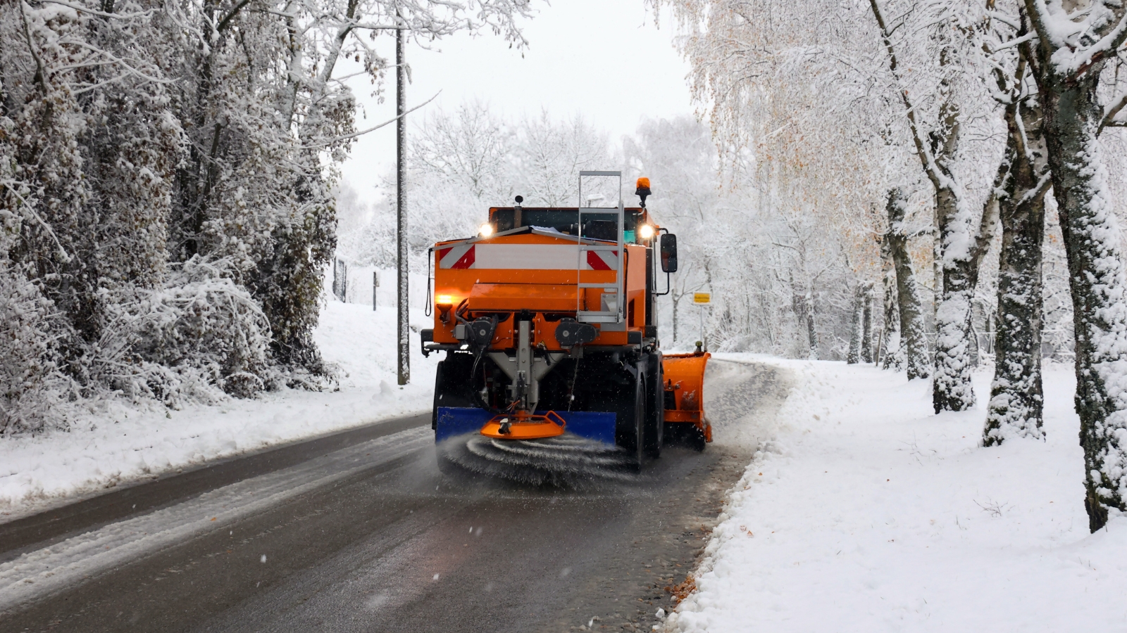 A large orange truck drives along a snowy road surrounded by trees and bushes, scattering salt.